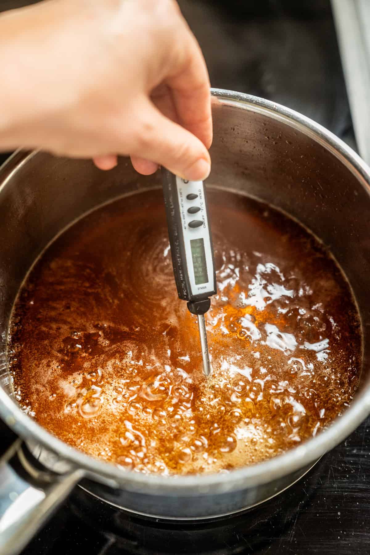 A hand holds a digital thermometer in a pot of bubbling brown liquid on a stovetop.