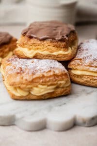 Three belgian cremekoeken filled with cream, topped with either chocolate icing or powdered sugar, sitting on a white marble board.