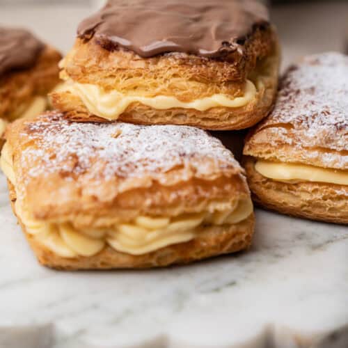 Three belgian cremekoeken filled with cream, topped with either chocolate icing or powdered sugar, sitting on a white marble board.