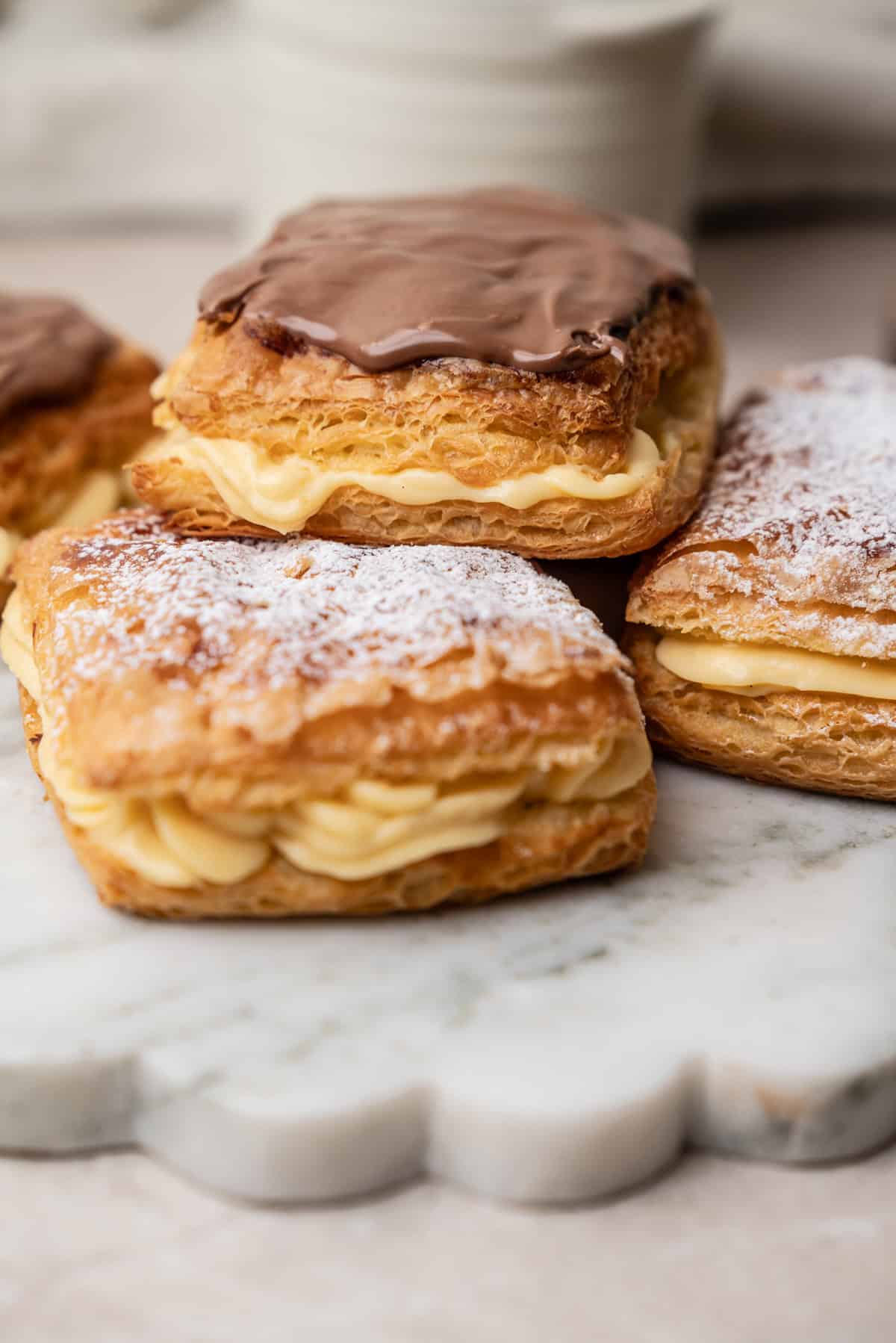 Three belgian cremekoeken filled with cream, topped with either chocolate icing or powdered sugar, sitting on a white marble board.