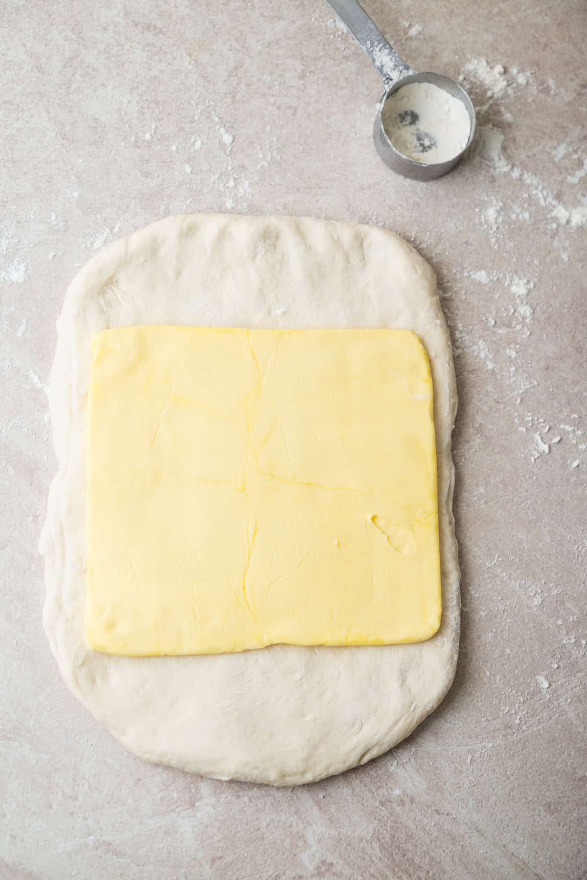 A square slab of butter placed on top of a rolled-out sheet of dough on a floured surface.
