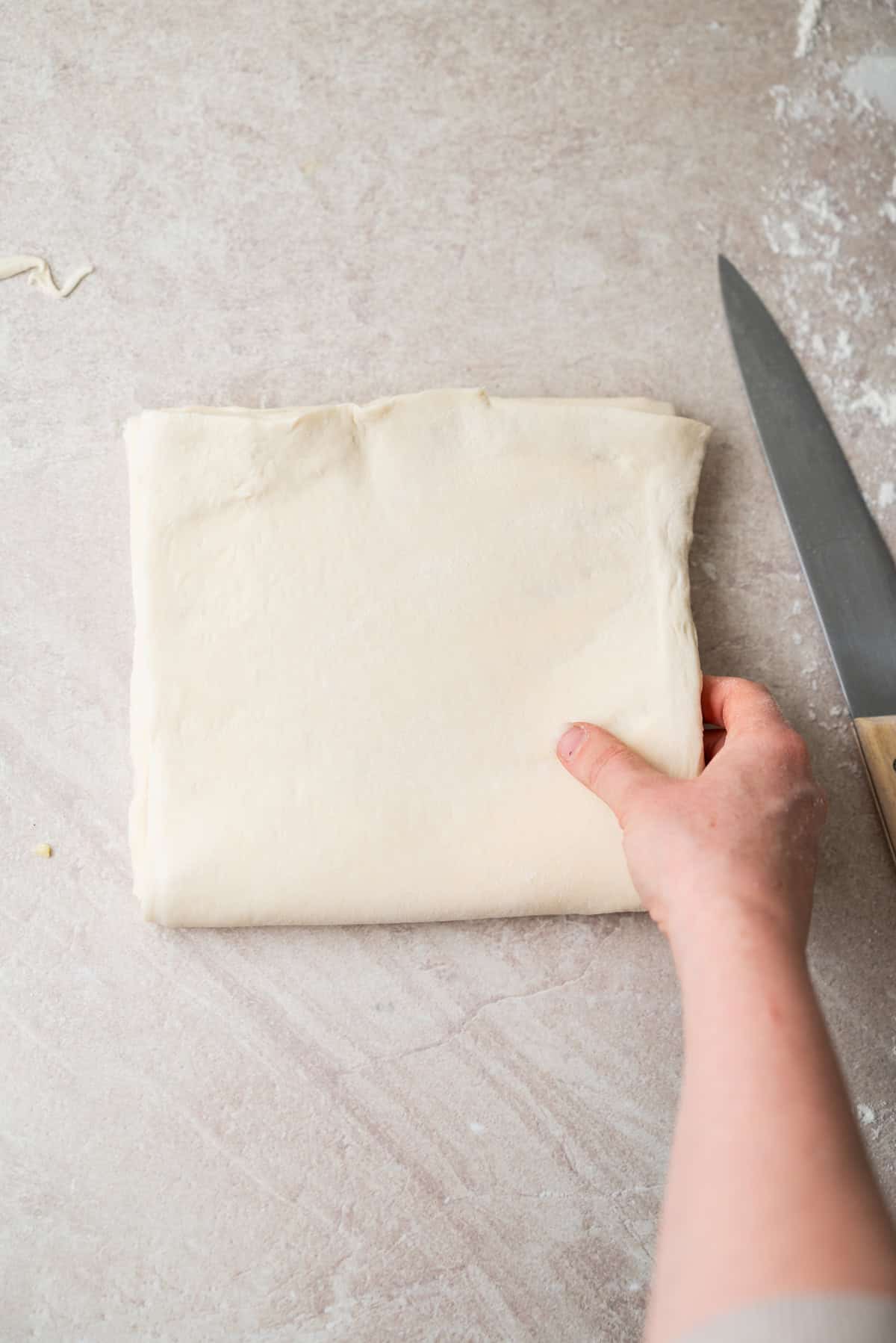 A hand holding a folded square of puff pastry dough next to a knife on a floured surface.