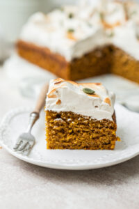 A slice of pumpkin cake topped with toasted meringue and a pumpkin seed on a white plate with a small fork.
