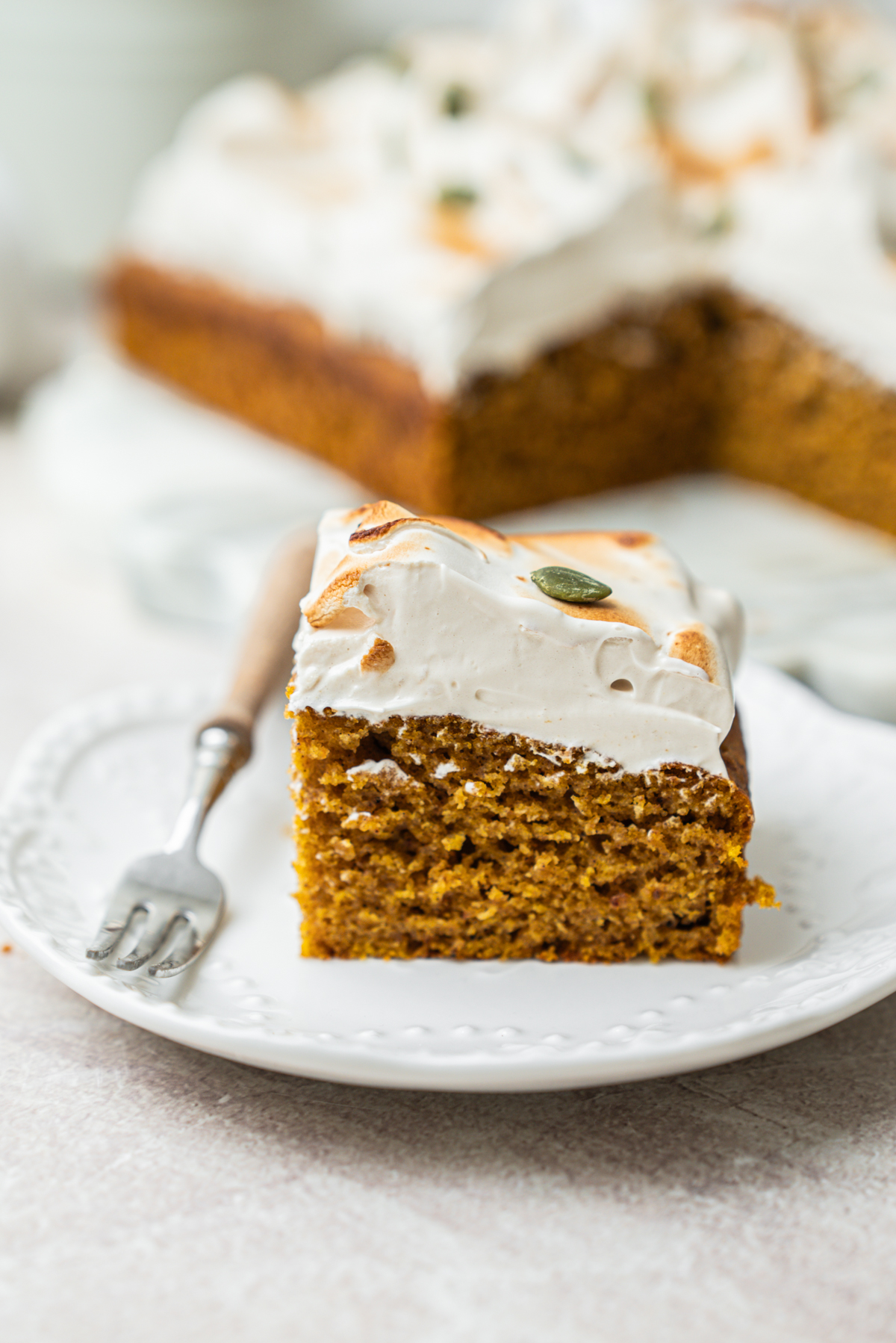A slice of pumpkin cake topped with toasted meringue and a pumpkin seed on a white plate with a small fork.