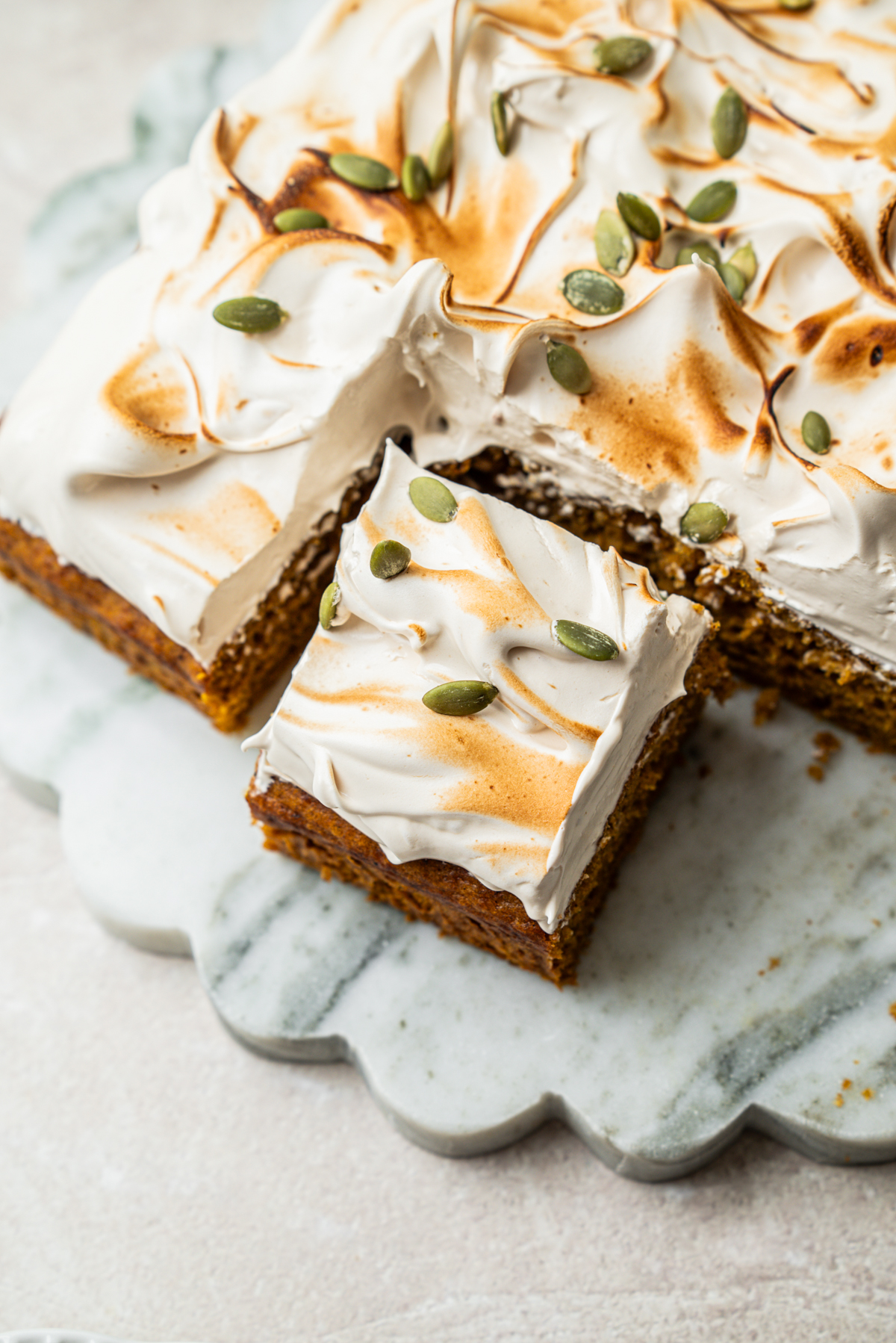 A square piece of spiced cake topped with toasted meringue and pumpkin seeds, resting on a marble serving board.