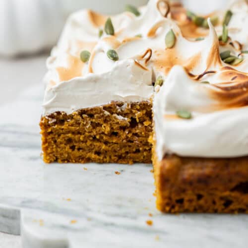 A slice of pumpkin cake topped with toasted meringue and pepitas on a marble platter.