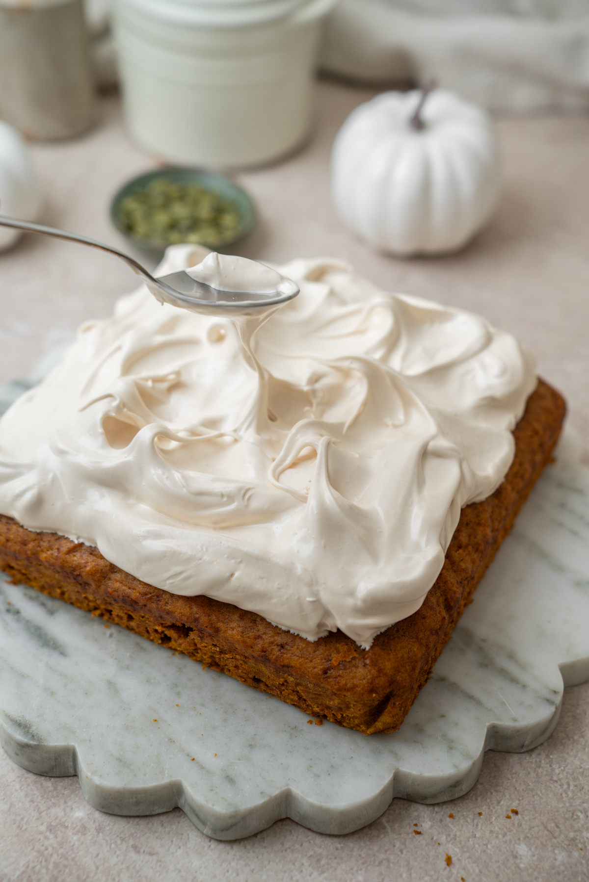 A spoon drizzles frosting onto a square cake with a thick layer of whipped cream topping.