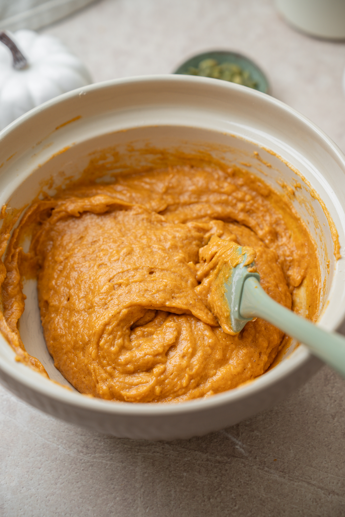 A rubber spatula sits in a mixing bowl filled with orange pumpkin batter.