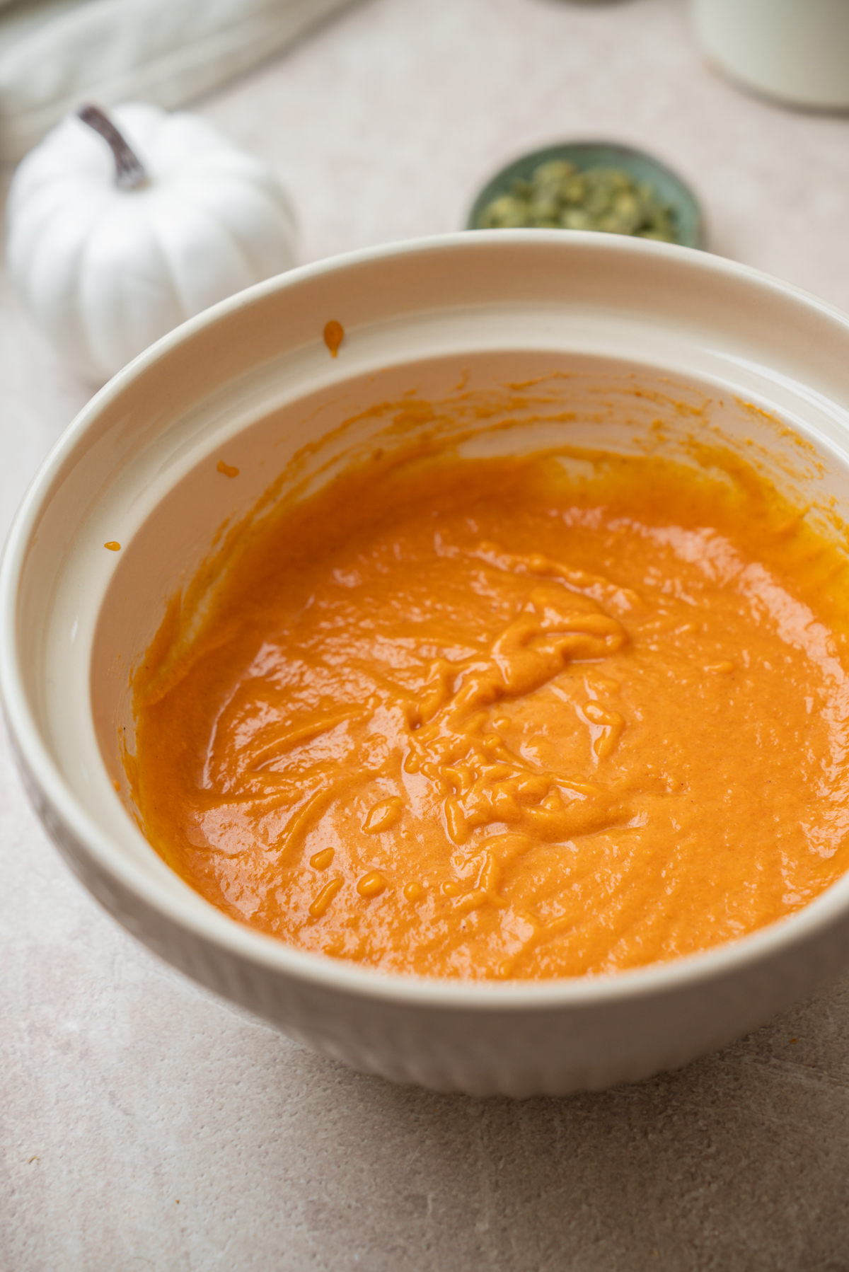 A large mixing bowl filled with vibrant orange pumpkin batter, with a small white pumpkin and a dish of seeds in the background.