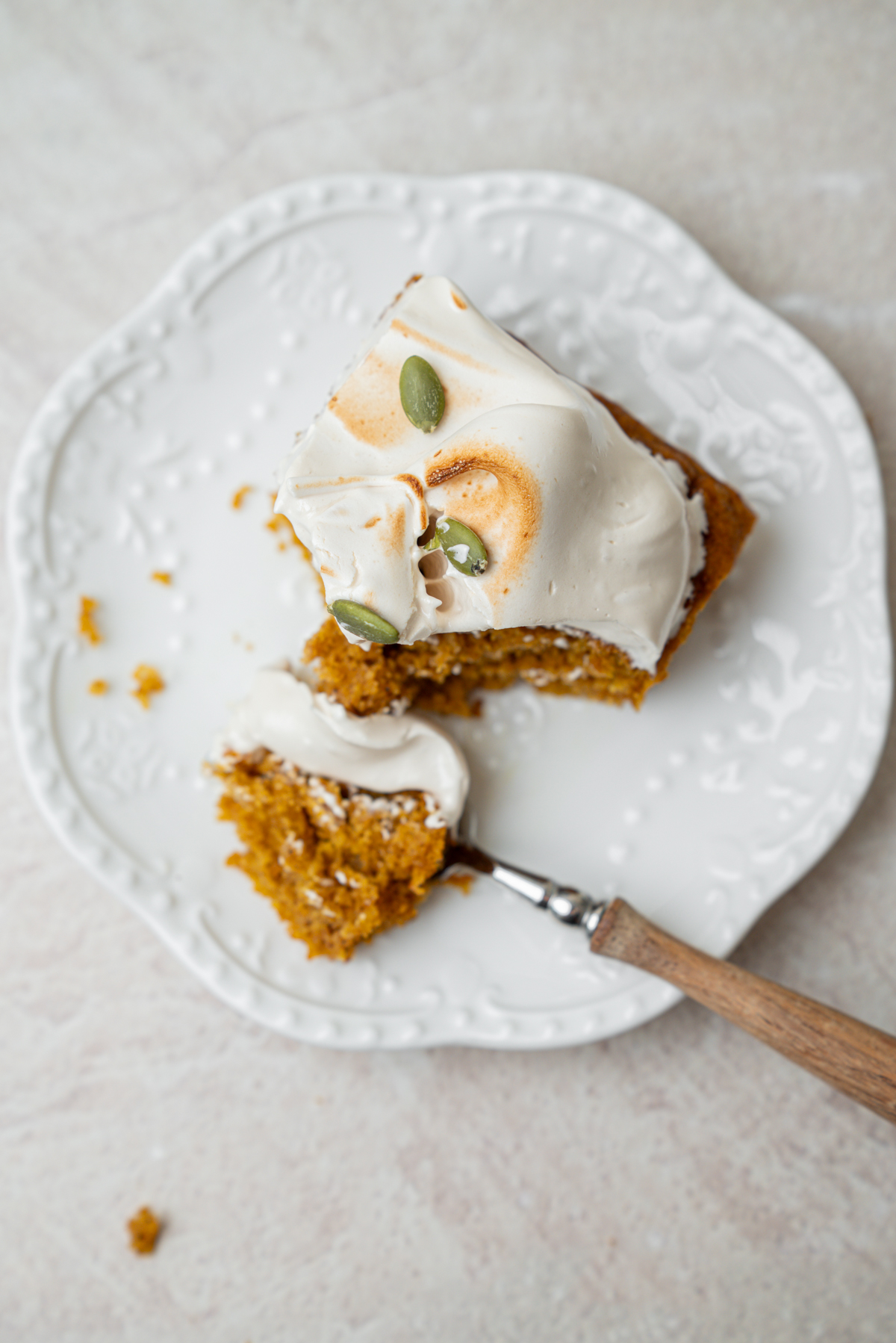 A slice of pumpkin cake topped with toasted meringue and pepitas on a decorative white plate, with a bite taken on a small spoon.