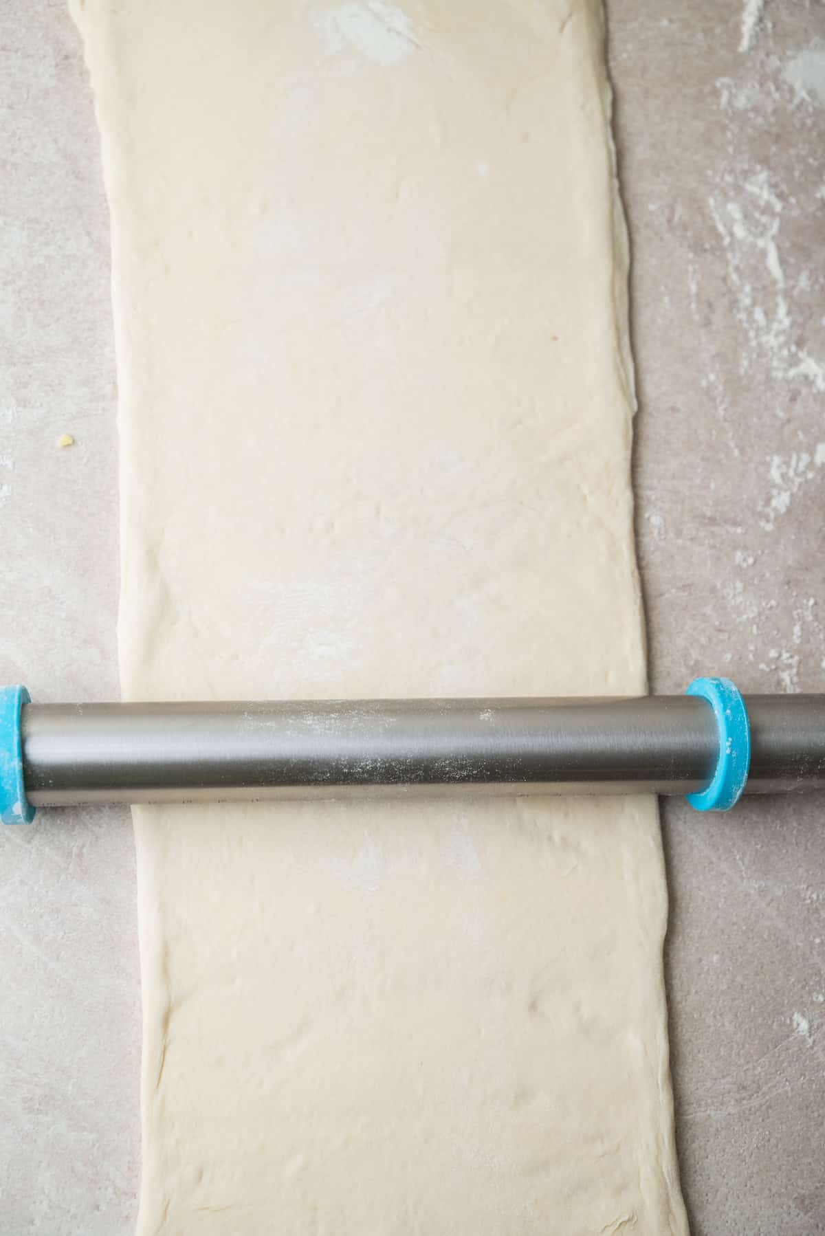 A metal rolling pin rolls out a rectangular sheet of dough on a floured surface.