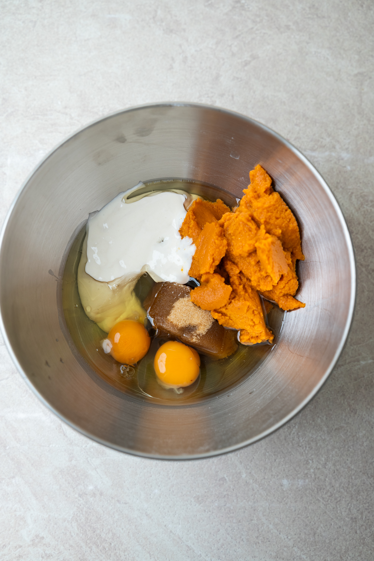 A metal mixing bowl containing pumpkin puree, two raw eggs, sourdough starter, and brown sugar.