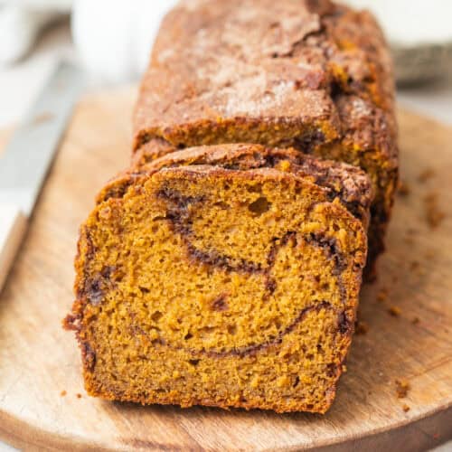 A loaf of pumpkin bread with a cinnamon swirl, sliced on a wooden cutting board.