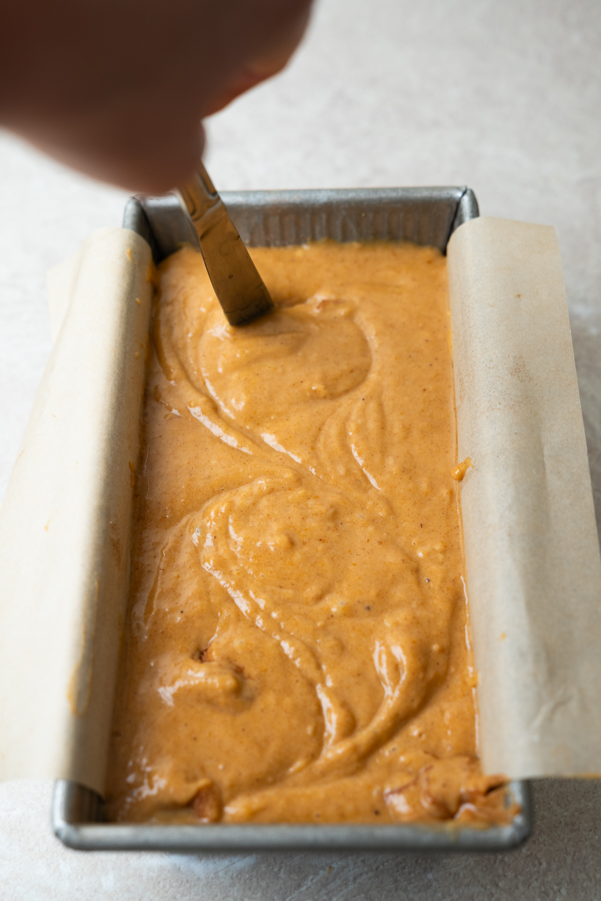 A metal knife smooths orange cake batter in a parchment-lined loaf pan.