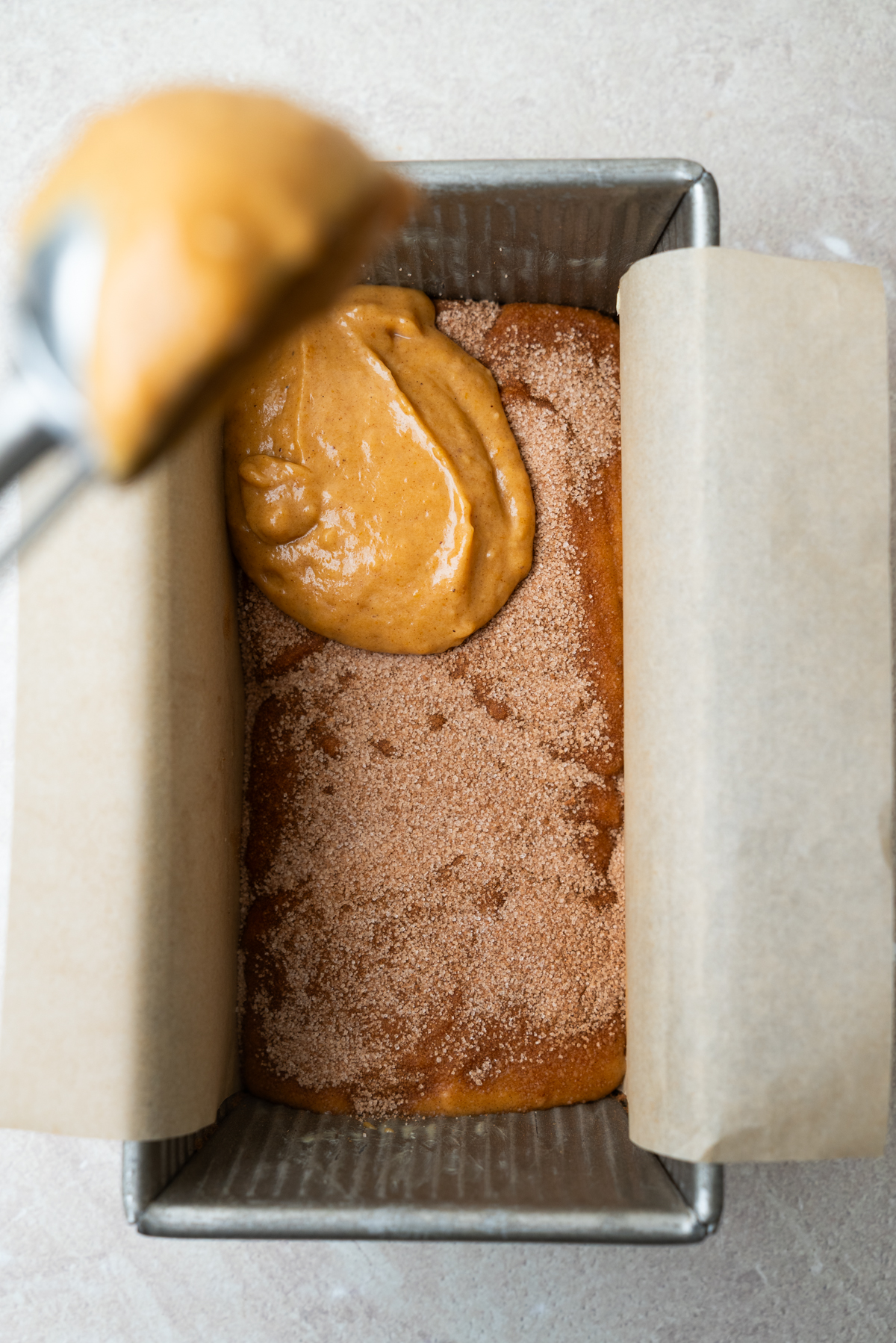 A metal scoop adds batter to a parchment-lined loaf pan containing a layer of batter topped with cinnamon sugar.