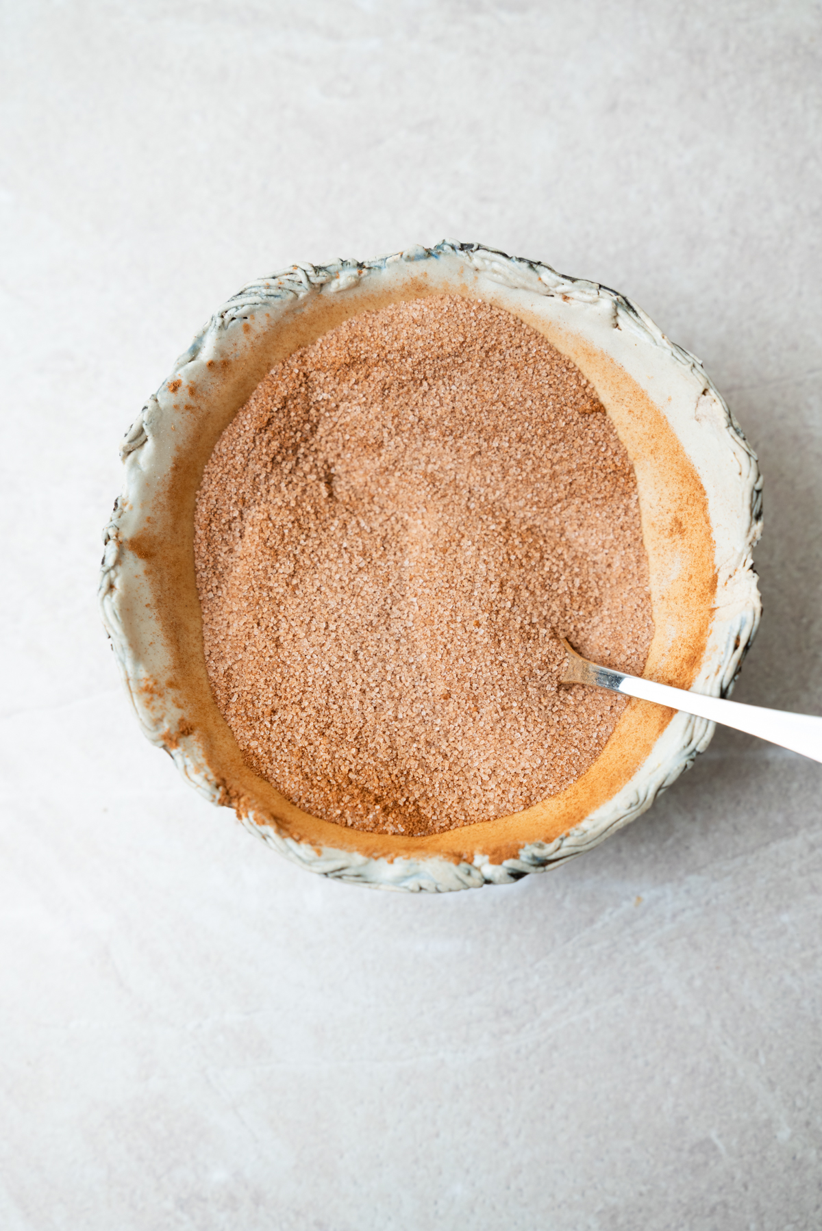 A small ceramic bowl filled with a brown cinnamon-sugar mixture and a silver spoon.