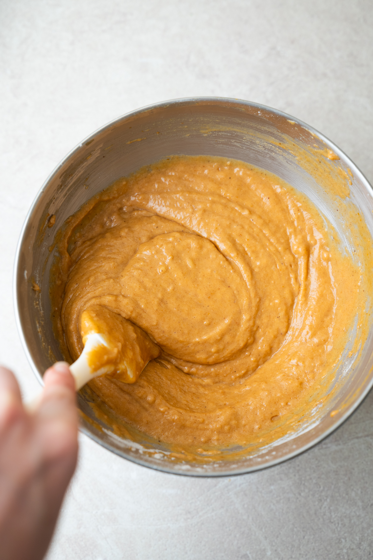 A hand uses a white spatula to stir thick, orange-colored batter in a metal mixing bowl.