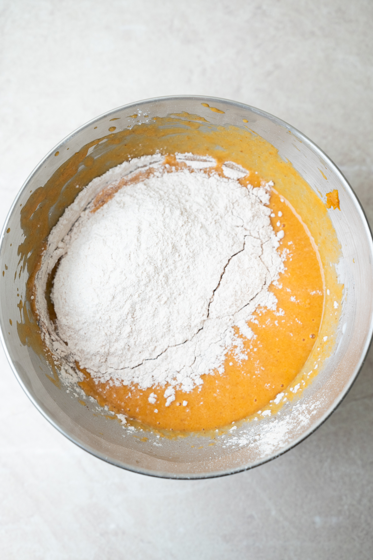 A mound of white flour sits in a metal mixing bowl atop orange cake batter.